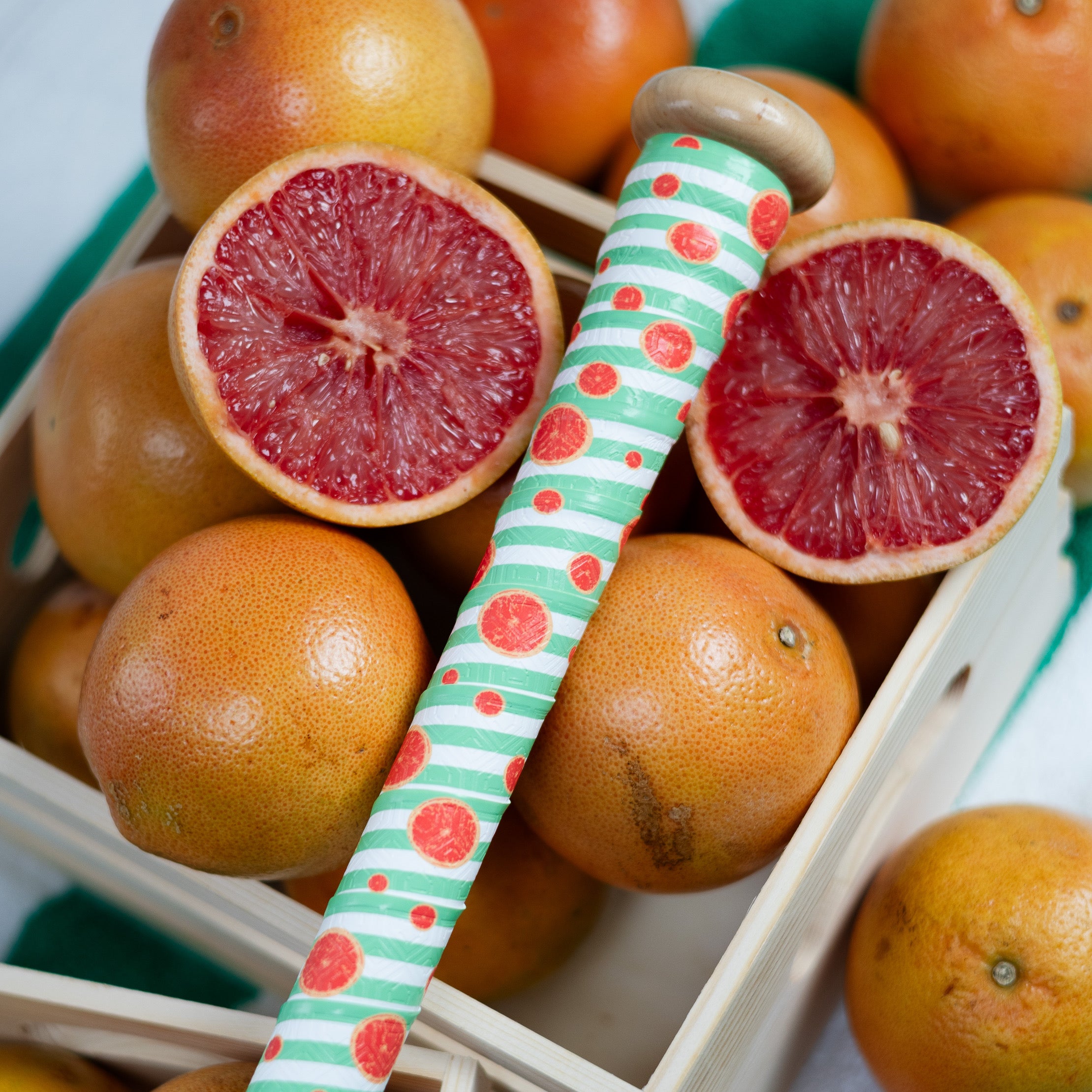 Grapefruits halved one showing red flesh, placed in a basket with a decorative straw with grapefruit bat grip on top of the basket, bat grip has light green stripes and grapefruit cross sections placed randomly throughout the bat grip.