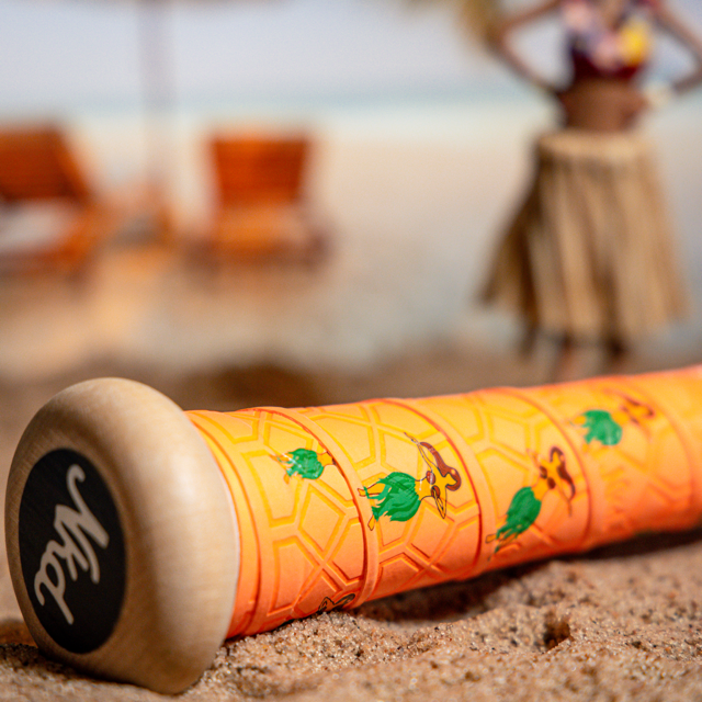 Image of the hulu girl bat grip wrapped around a wooden bat laying in the sand with a hulu girl statue out of focus in the background.