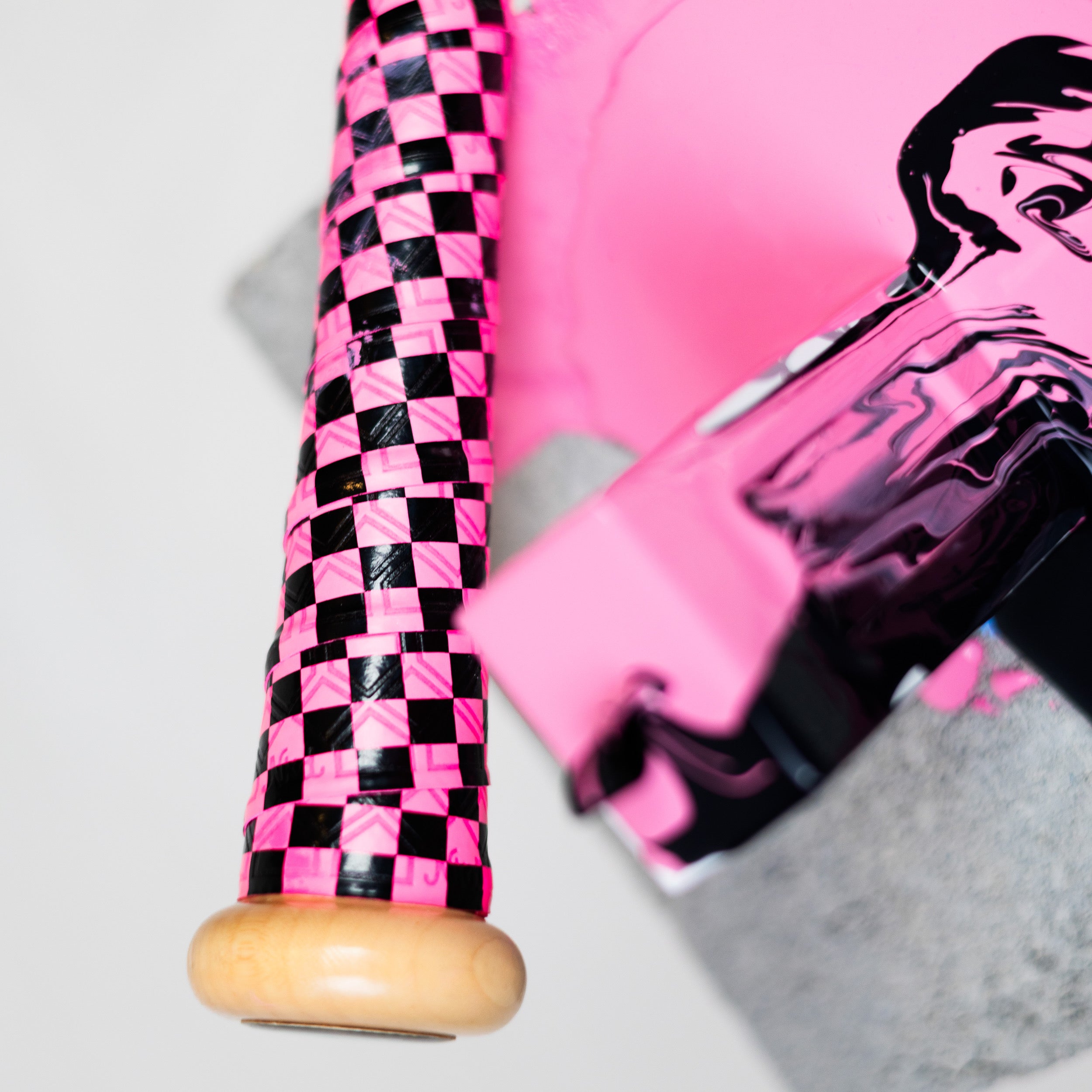 Close-up shot of baseball bat handle wrapped with bright pink and black checkerboard grip. Next to grip is spilled and mixed pink and black paint.