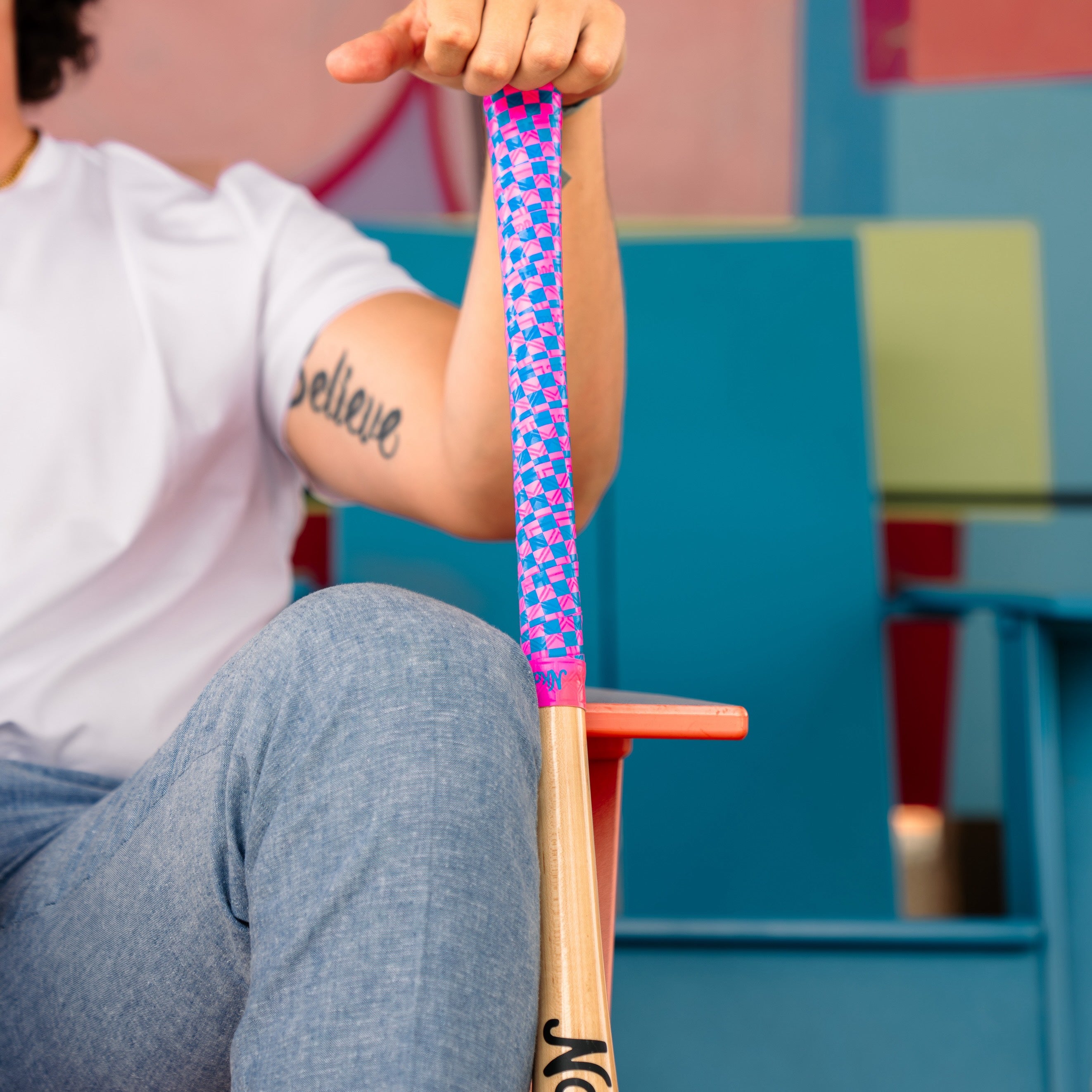 Photo of a man sitting down propping up a wooden bat with a pink and blue checkerboard bat grip.