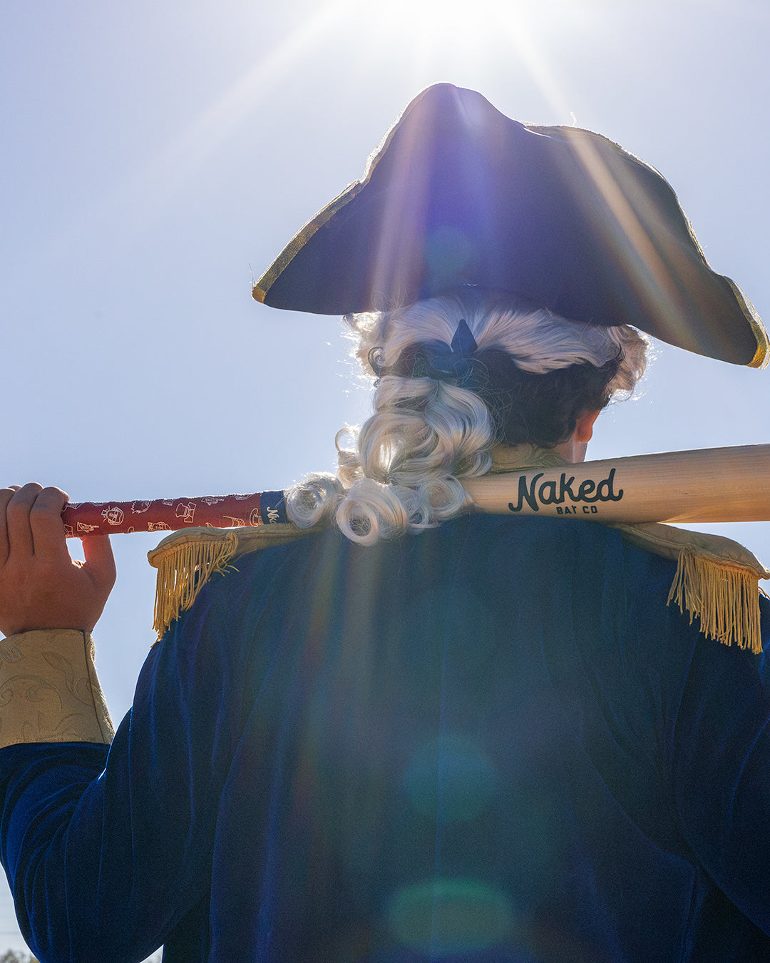 Back of person in George Washington costume holding wood bat over back of shoulders with a red USA 250 bat grip wrapped on the bat.