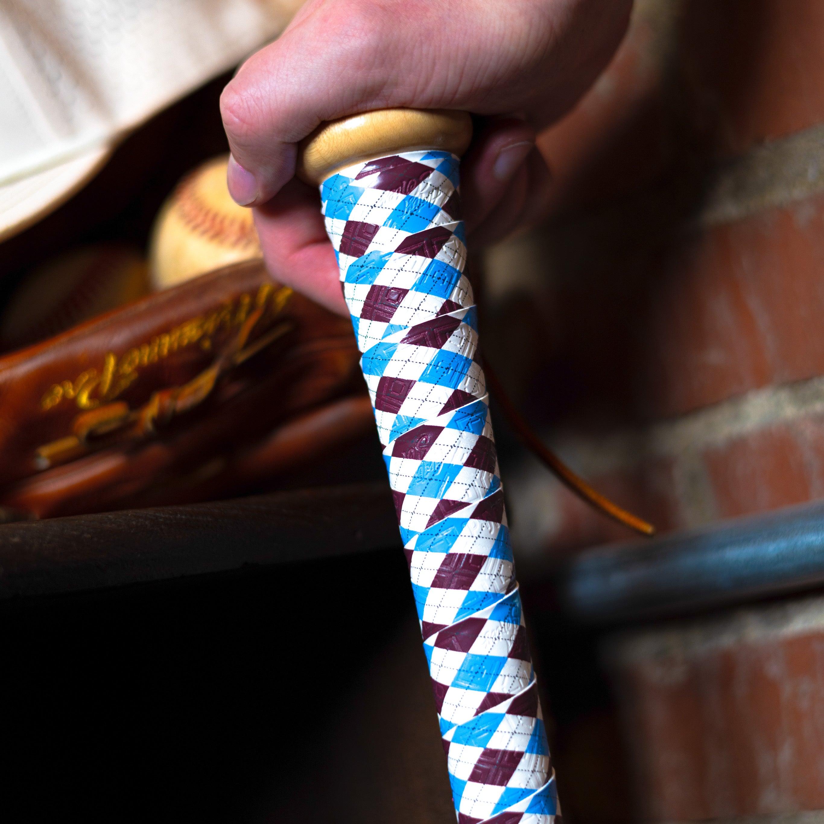 Hand holding a baseball bat handle with the bat handle wrapped with a powder blue and maroon argyle bat grip. A ball and glove sit behind the photo subject with a brick wall background.