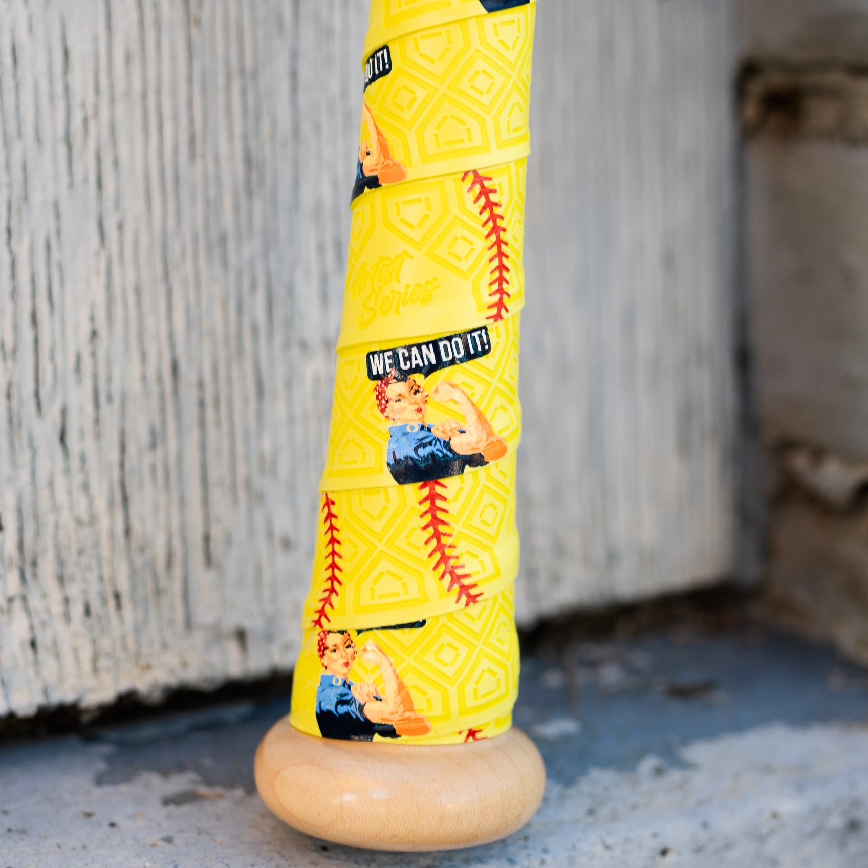 A baseball bat handle wrapped with a yellow Rosie the Riveter themed bat grip with softball seams intermingled on the grip. Bat handle sits in front of a weathered wood backdrop.
