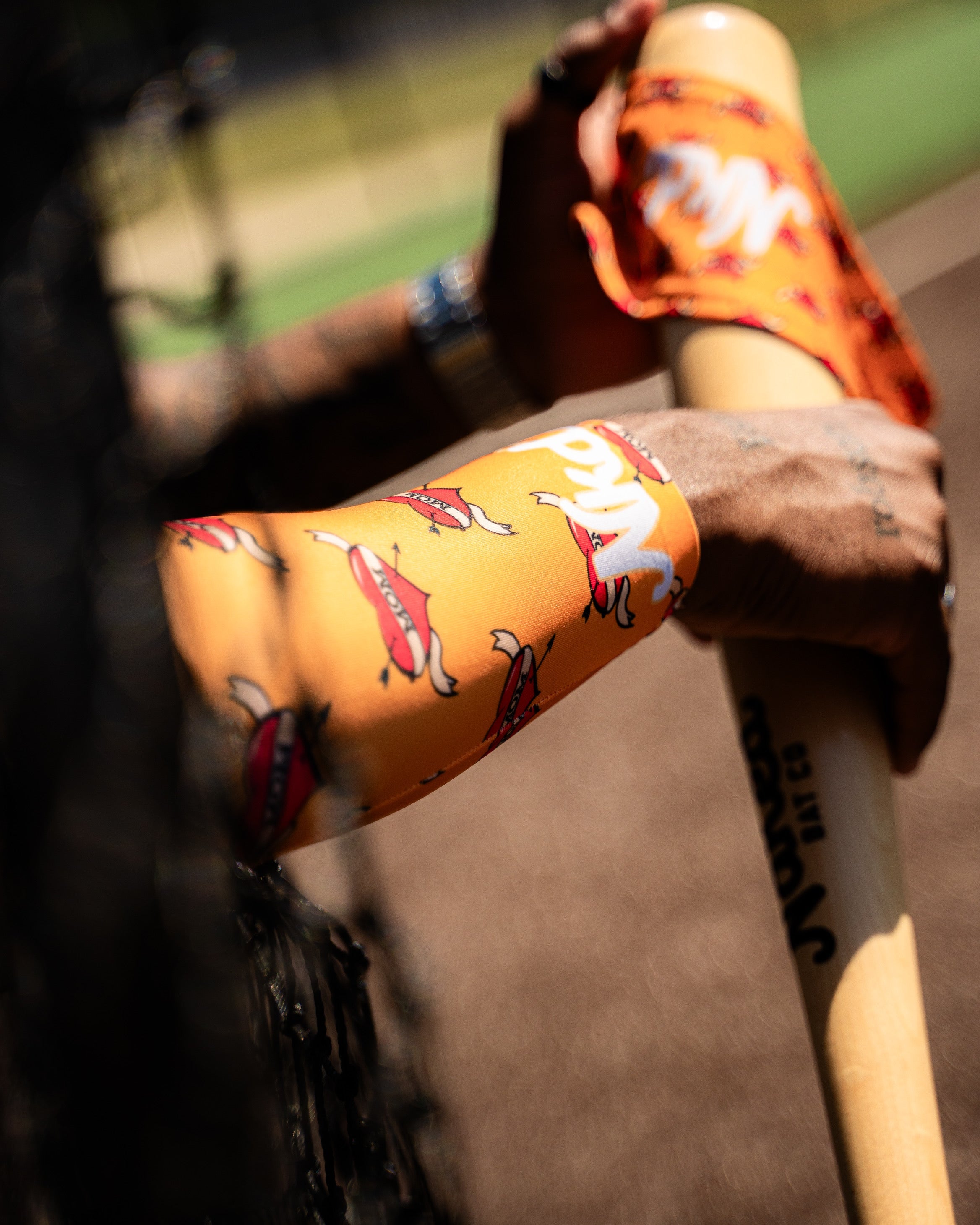 photo of man's arm wearing an orange arm sleeve with a mom tattoo with a heart on it. arm is holding wood baseball bat with the matching mom headband on the top of the bat. background baseball field.