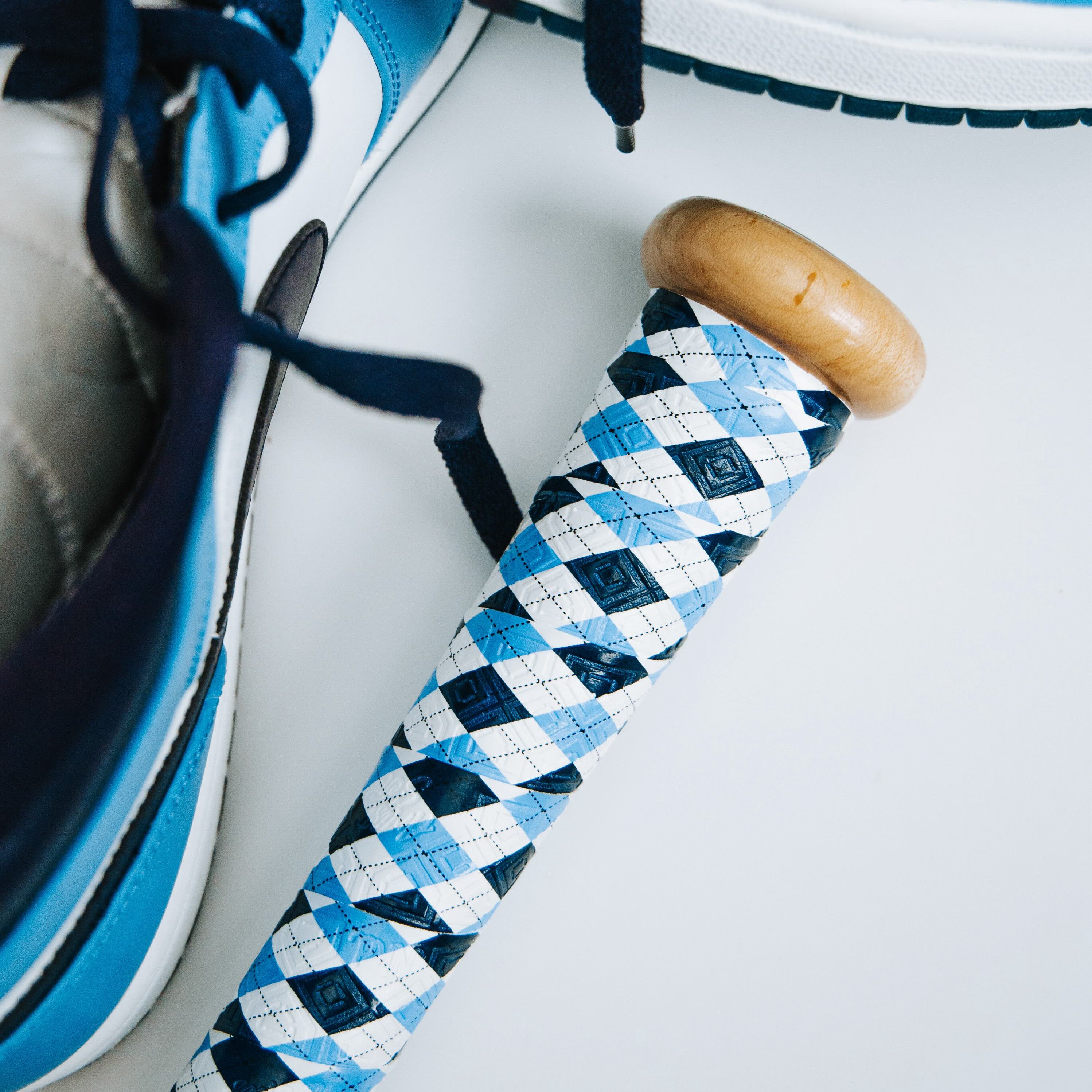 A photo of the blue and dark blue plaid bat grip installed on a wooden bat with shoes in the background of the photo that are also blue and dark blue. 