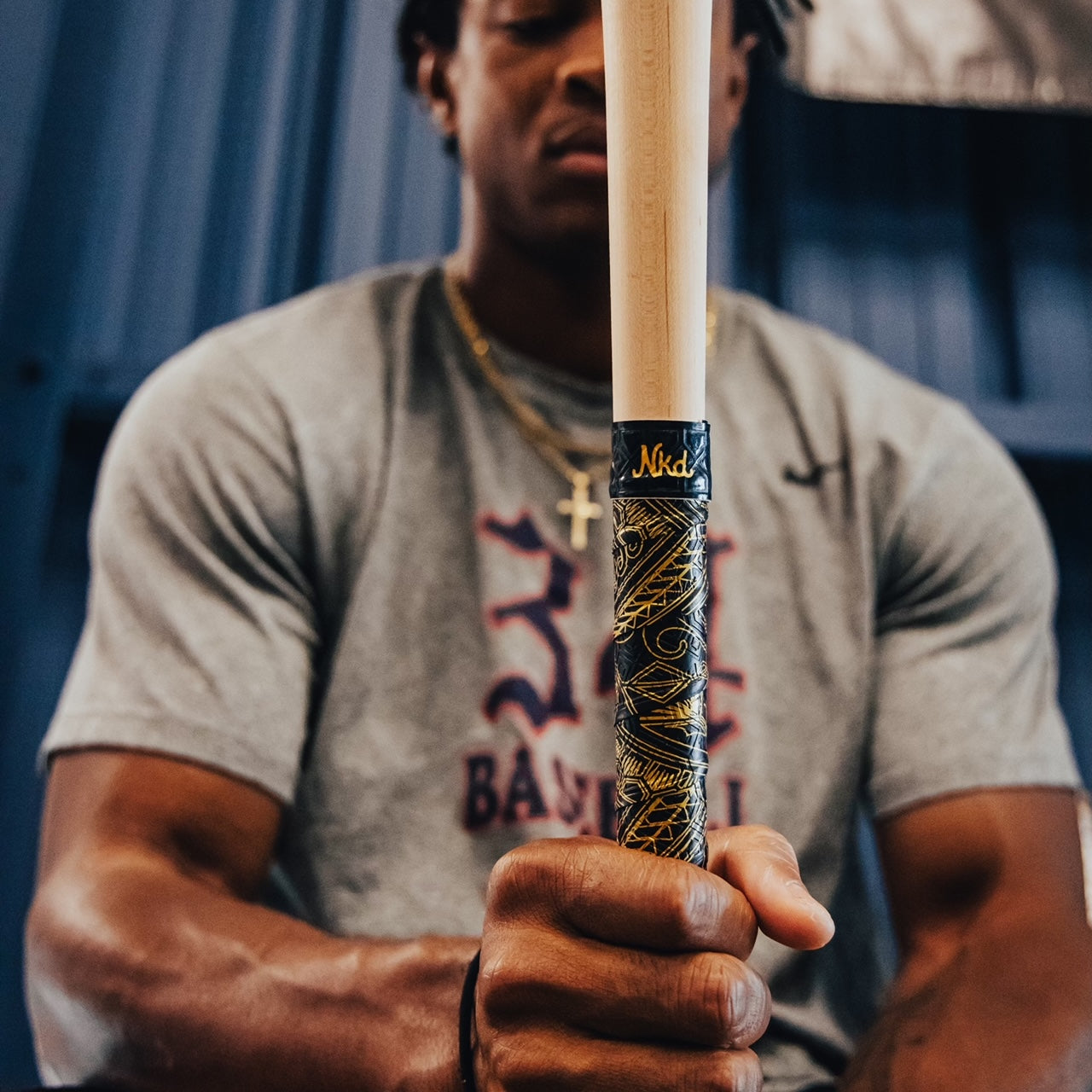 Photo of the golden tribal bat grip wrapped around a wooden bat being held upright by a man in a grey t-shirt