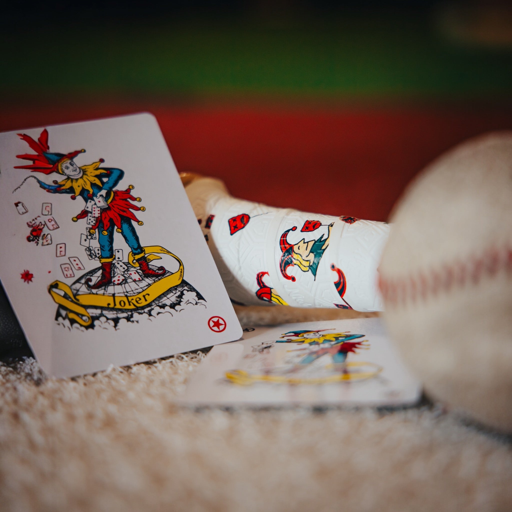 Photo of the joker bat grip laying on the ground with a Joker playing card and baseball in the frame. 