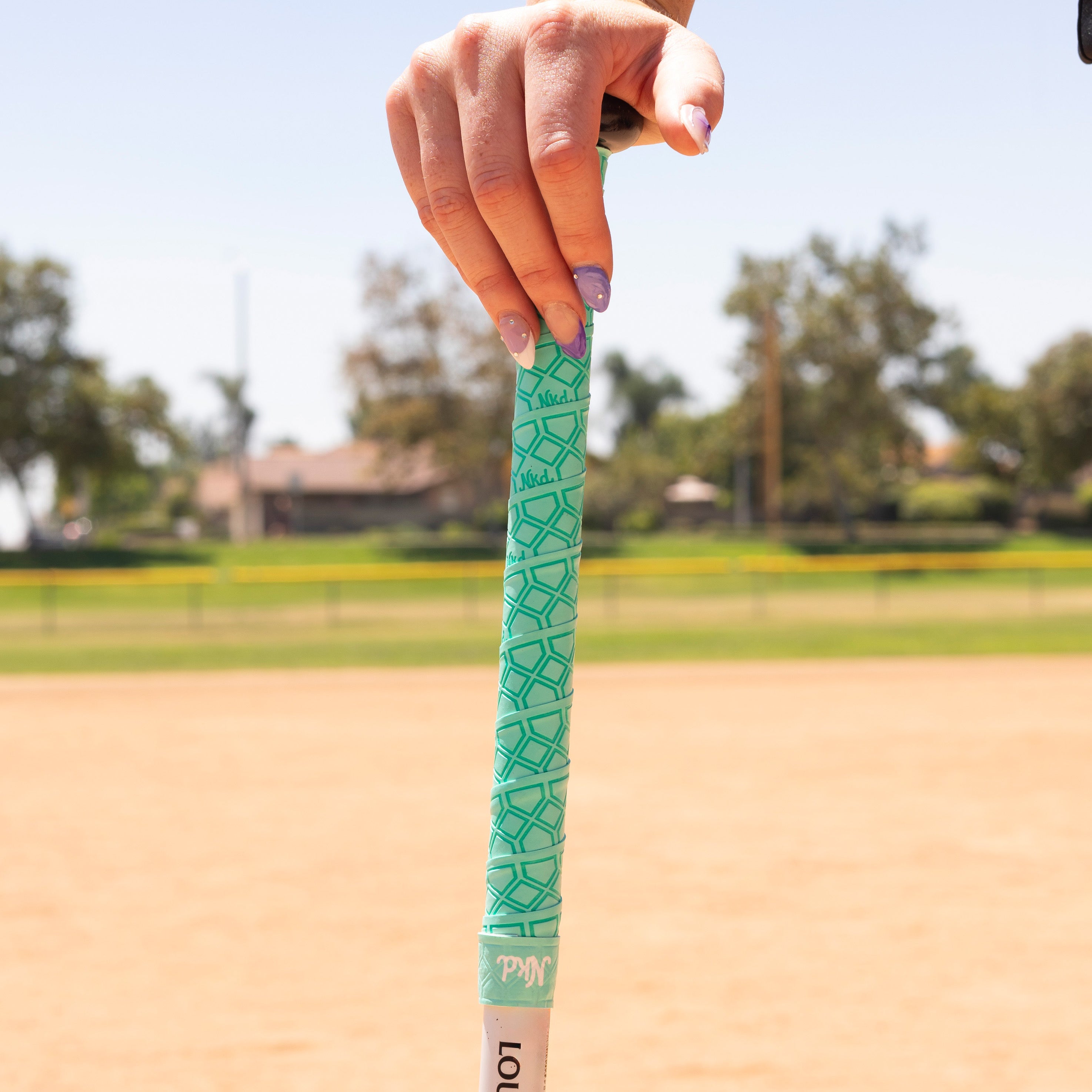 Photo of a hand leaning on a wooden bat wrapped with the mint colored bat grip on a baseball field. 