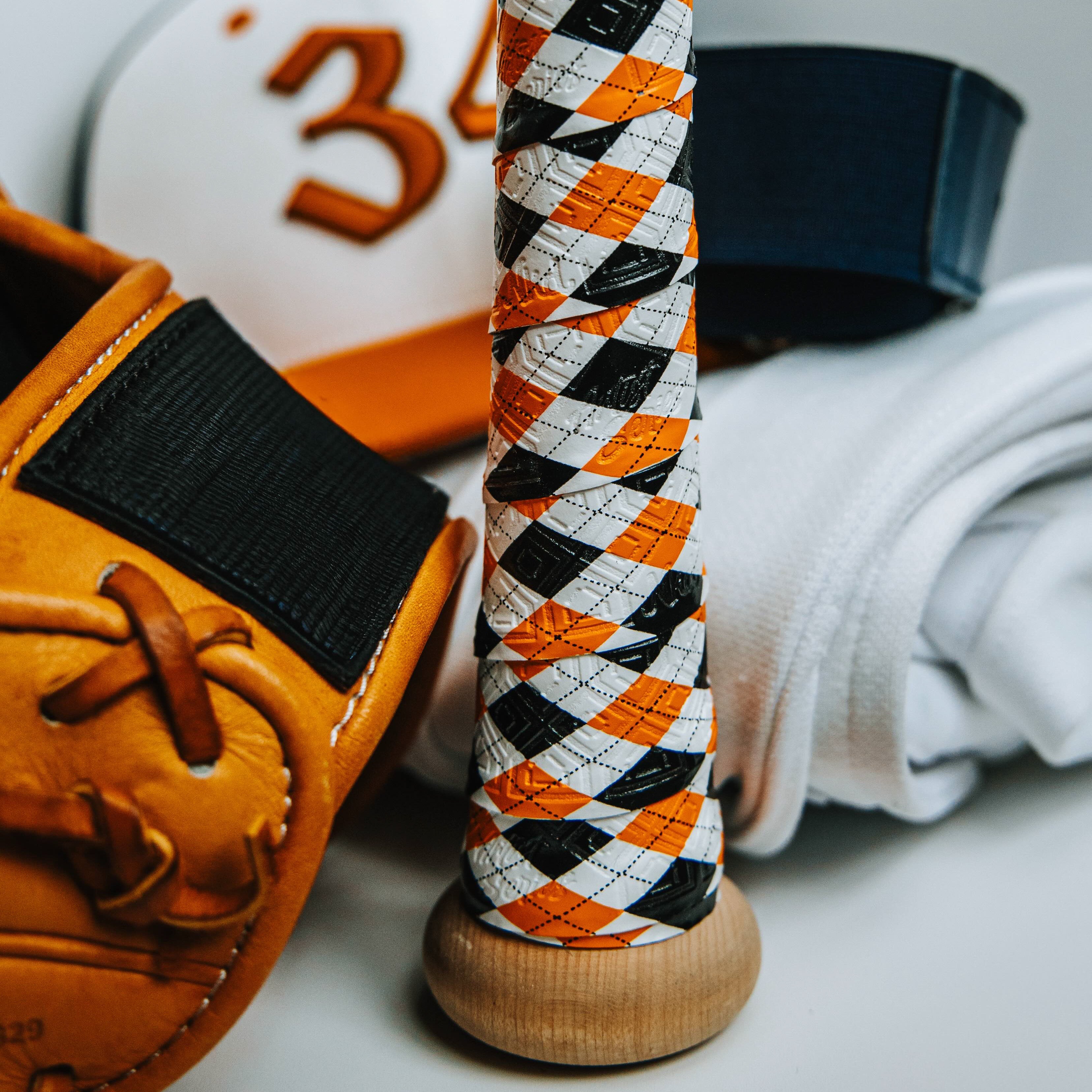 Photo of the Orange and Black plaid bat grip on a wooden bat with miscellaneous batting gear in the background. 