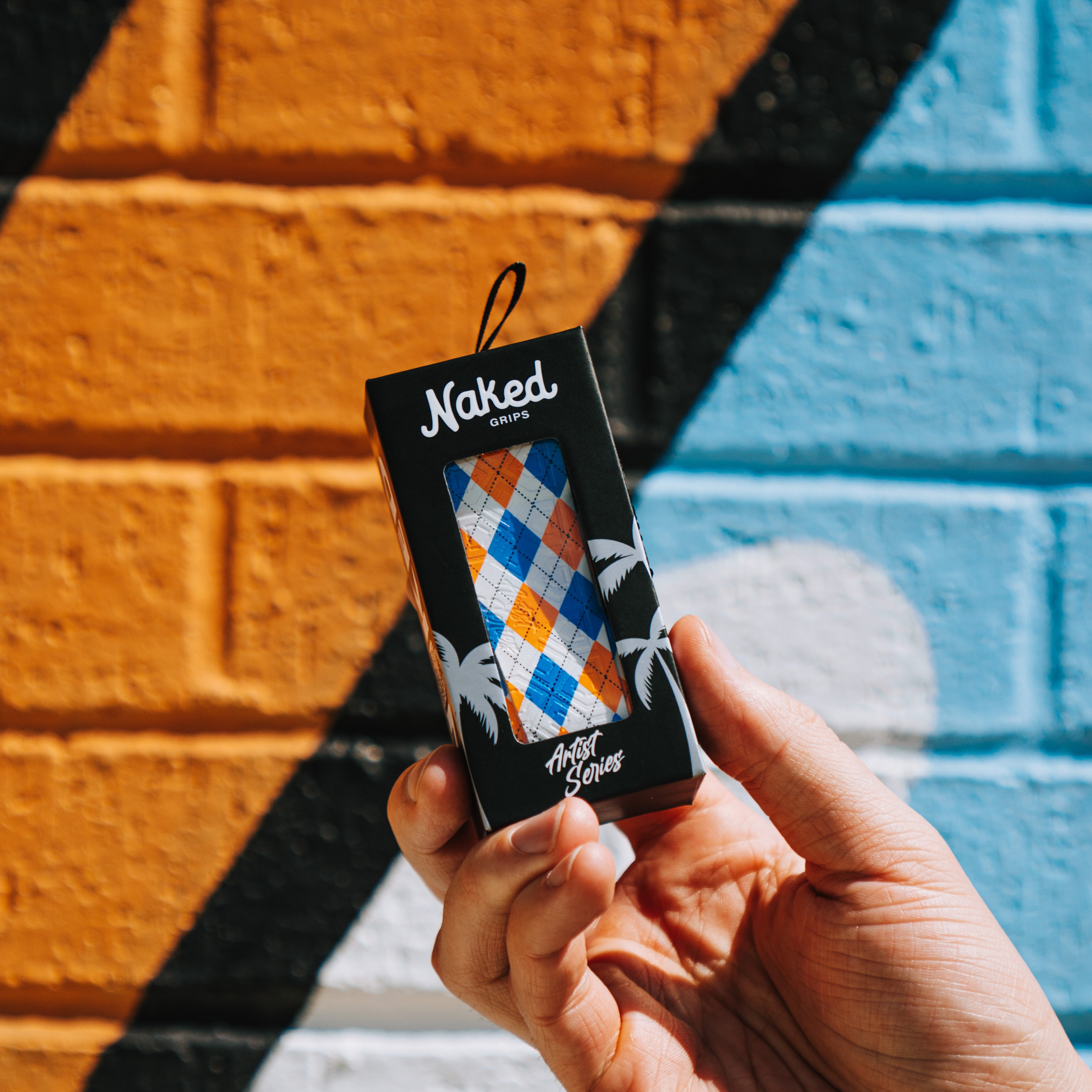 A photo of the naked grips orange and blue plaid grip being held in someones right hand with an orange and blue background. 