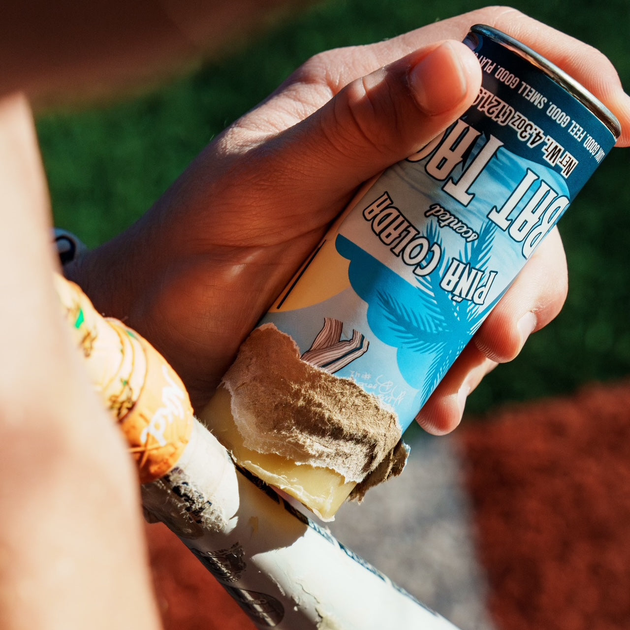 Photo of the Pina Colada scented bat tack being applied to a wooden bat with green grass and dirt in the background. 