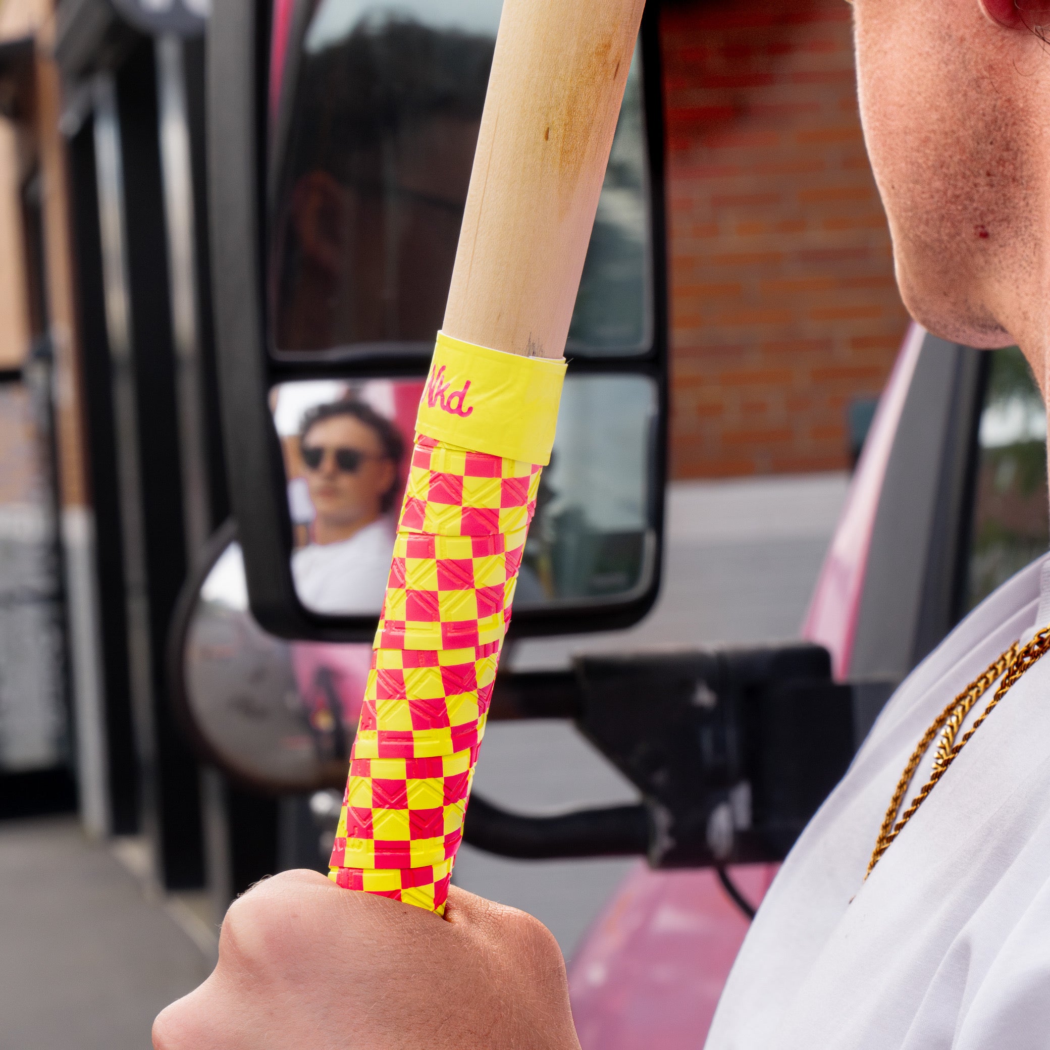 Photo of a man looking in a truck mirror holding a wooden bat wrapped with the pink and yellow checkerboard bat grip. 