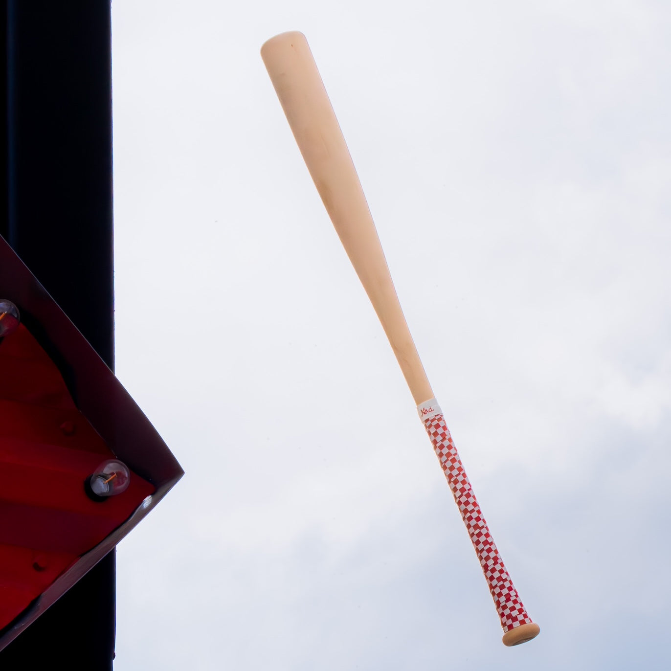Photo of a wooden bat on a white background wrapped with a red and white checkerboard grip. 