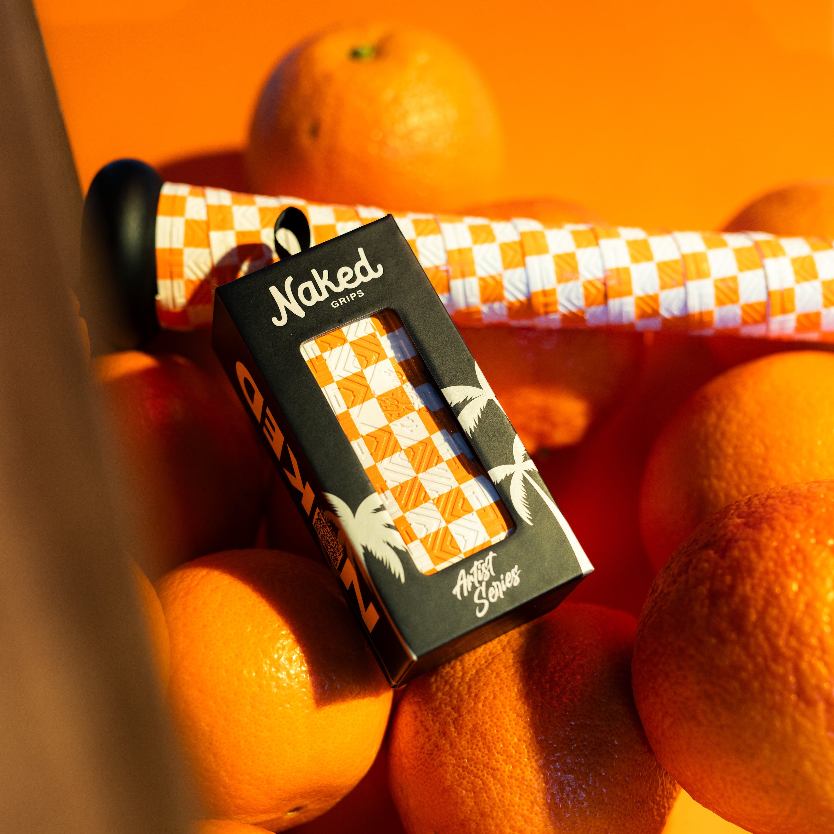 Black artist series grip box with Tennessee themed orange and white bat grip inside. Behind package are oranges on an orange backdrop with a baseball bat handle wrapped with the same bat grip. 