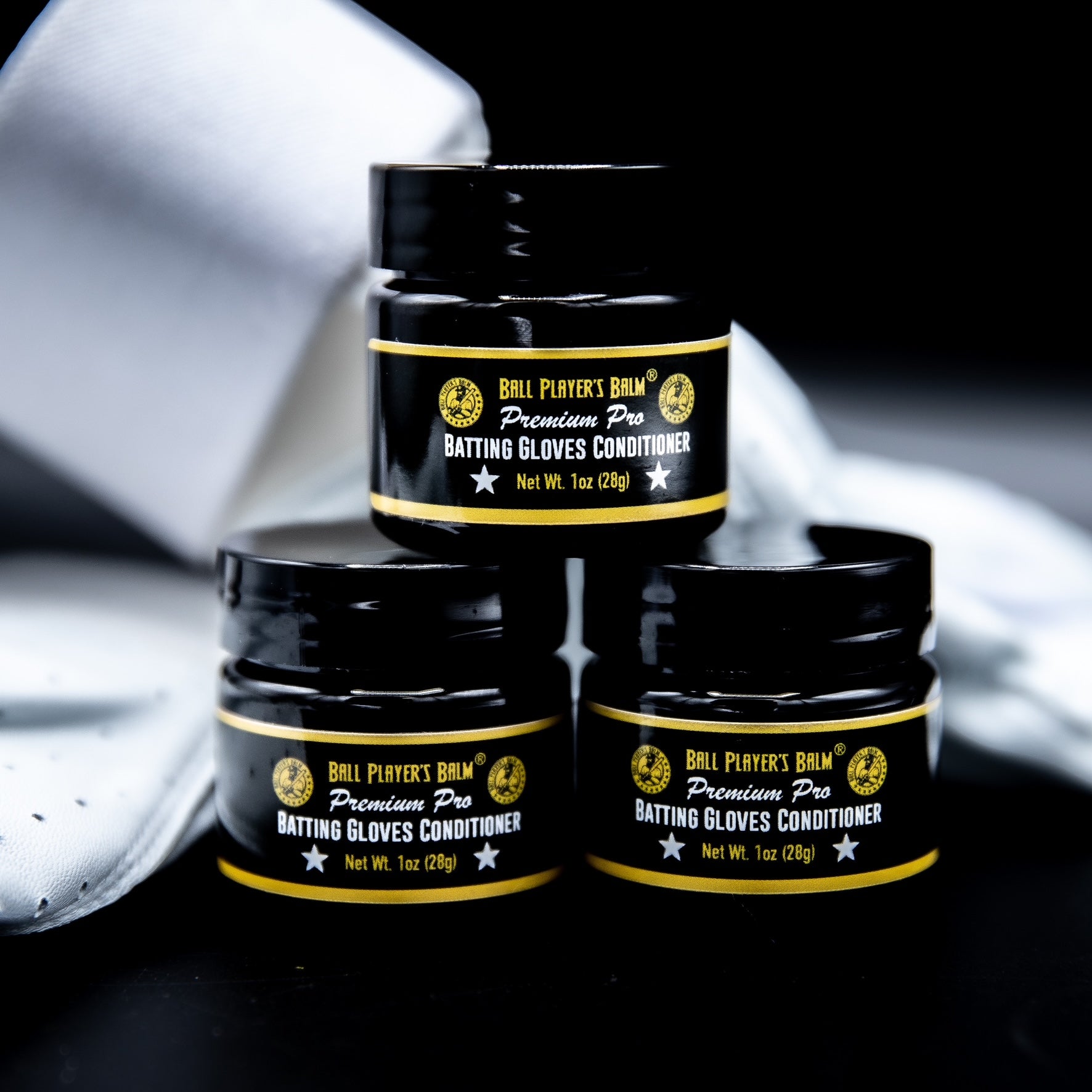 photo of triangular stack of three black one ounce containers of batting glove conditioner. containers are closed. white batting gloves are backdrop of photo.