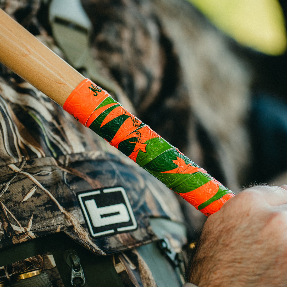zoomed in shot of man in camo gear holding orange and green camo  hunting themed bat grip with deer and duck outlines in the grip design.