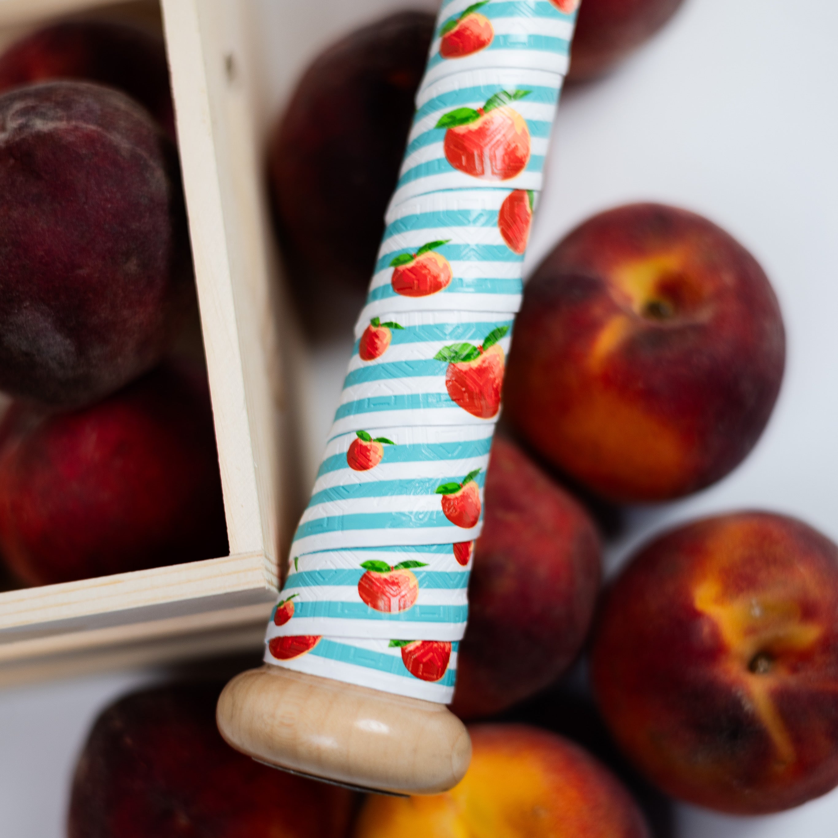 peach theme bat grip on a baseball bat handle with peaches interspersed throughout the grip which also features a turquoise and white striped pattern. Behind the bat handle sits actual peaches.