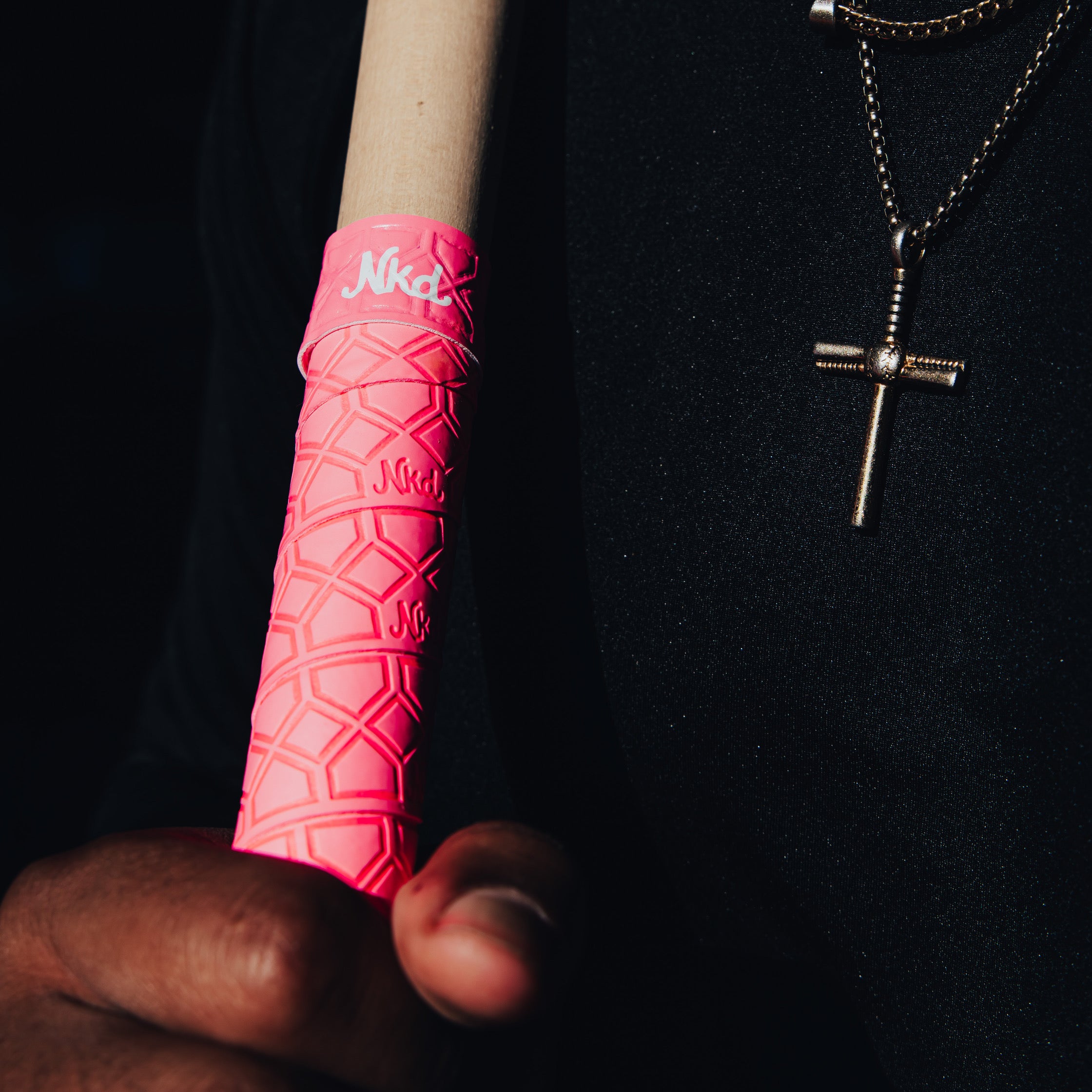 Photo of the naked Grips pink bat grip being held upright close to someones chest with a cross hanging around the persons neck. 
