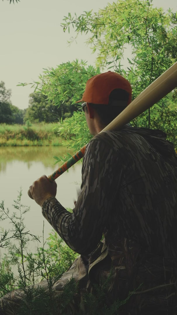 man in hunting gear holding baseball bat with naked grips ducks and bucks hunting themed bat grip. grip has orange and green camo and duck and deer outlines for the design.